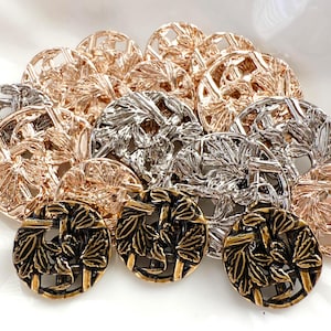 May include: A collection of round metal buttons featuring an openwork design with embossed leaf patterns. The buttons are finished in metallic rose gold, silver, and antique bronze tones, arranged in a cluster on a plain white surface.