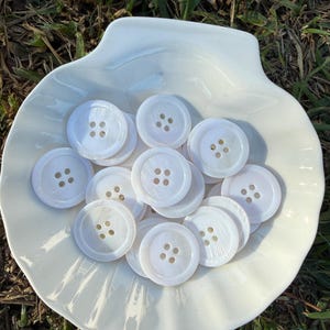 May include: A collection of white, round buttons with four holes, resting in a white, seashell-shaped dish. The buttons have a pearlescent sheen and are arranged on a textured surface, suggesting a craft or sewing project.