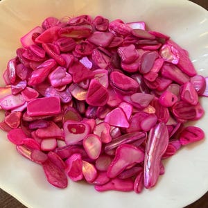 May include: A collection of pink and magenta shell chips fills a white, scalloped bowl. The chips vary in size and shape, with iridescent surfaces reflecting light. The bowl sits on a dark brown surface, creating a contrast with the bright colors of the shells.