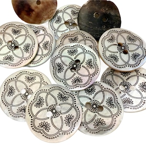 May include: A collection of round, off-white buttons with a floral design in black ink. Each button has a four-petal flower in the center, surrounded by a dotted border. Two larger, darker buttons are also visible.
