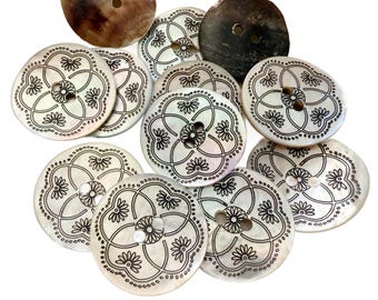 May include: A collection of round, off-white buttons with a floral design in black ink. Each button has a four-petal flower in the center, surrounded by a dotted border. Two larger, darker buttons are also visible.