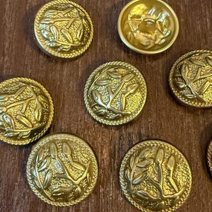 May include: A collection of gold-colored metal buttons. Most buttons feature a detailed relief design of leaves and stems. One button has a different design. The buttons are round with a textured edge.