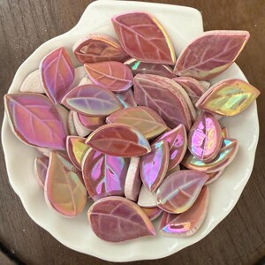 May include: A collection of iridescent, leaf-shaped mosaic tiles in shades of pink, purple, and gold, displayed in a white, shell-shaped dish. The tiles have a glossy finish and are arranged to show their detailed veining.