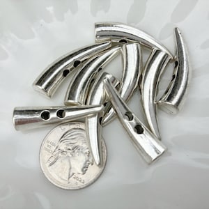 May include: A collection of silver-colored horn-shaped toggle clasps with three holes for sewing. A U.S. quarter coin is included for size reference. The clasps are arranged on a white, slightly textured surface.