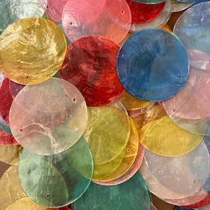 May include: A collection of colourful, translucent, round shell discs in shades of pink, red, yellow, green, and blue. Each disc has a small hole near the edge. The discs are scattered, overlapping each other.
