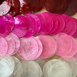 May include: A collection of circular, flat shell discs arranged in rows by colour, transitioning from deep red at the top to bright pink, light pink, and translucent white at the bottom. Each disc features a small, pre-drilled hole near the edge.