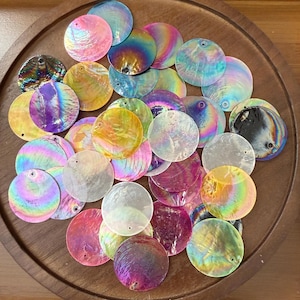 以下が含まれることがあります： ピンク、青、黄色、紫など、さまざまな色の虹色の丸い貝殻ビーズのコレクション。各ビーズには、紐を通すための小さな穴があります。ビーズは木製のトレイに配置されています。