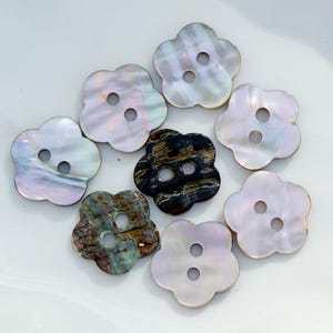May include: A collection of flower-shaped buttons with two holes each. The buttons have a pearlescent finish in shades of white, gray, and iridescent colors. One button is dark with black and brown marbling, and another is green and gray.