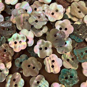 May include: A collection of flower-shaped buttons crafted from iridescent mother-of-pearl shell. Each button features two central holes and displays a variety of natural color tones, including soft pinks, creams, browns, and deep greens.