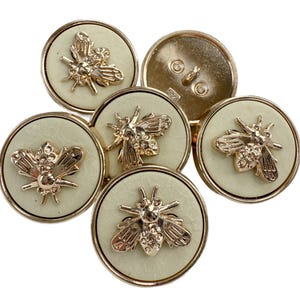 May include: A collection of round buttons with a light green center and gold-colored rims. Each button features a detailed gold bee design. The buttons are arranged in a cluster, showcasing their decorative appeal. Ideal for sewing and craft projects.