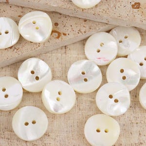 May include: A collection of round, white mother-of-pearl buttons, each featuring two central holes. These circular fasteners display a smooth, iridescent surface with natural variations in luster and tone, arranged in a scattered pattern.