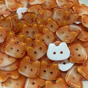 May include: A collection of cat-shaped buttons in translucent orange with red striped details and opaque white, each featuring two central holes for attachment. The buttons are arranged in a dense pile, showcasing their distinct feline silhouettes.