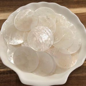 May include: A collection of circular, flat mother-of-pearl shell discs with a smooth, iridescent finish. Each disc features a small, centered hole for threading. The pieces are white with subtle, natural pearlescent variations in texture.