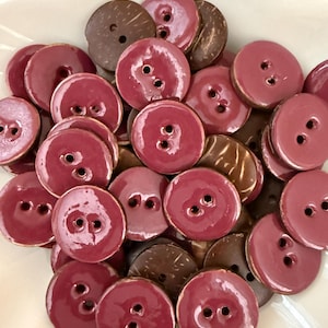 May include: A collection of round buttons in shades of burgundy and brown. The buttons have two holes for sewing and are arranged in a white, petal-shaped dish. Some buttons have a glossy finish, while others appear to be made of wood.