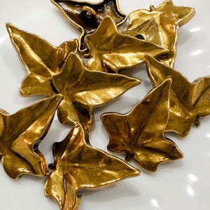 May include: A collection of gold-colored leaf-shaped decorative elements. The leaves have a detailed, textured surface and a metallic finish. The arrangement suggests a nature-inspired design, possibly for jewelry or craft projects.