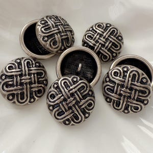 May include: A collection of silver-colored buttons with a detailed, interwoven design. The buttons have a round shape with a raised pattern and a black backing. These decorative buttons are ideal for sewing and craft projects.