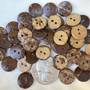 Può includere: Una collezione di bottoni rotondi a due fori realizzati con gusci di cocco naturali. I bottoni variano in tonalità di marrone e beige, con motivi di venature visibili. Una moneta è inclusa per riferimento alle dimensioni.