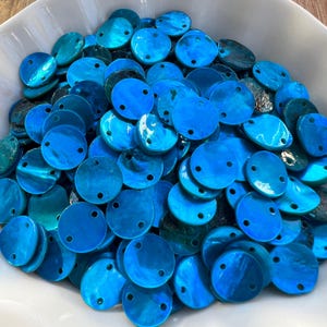 Puede incluir: Una colección de botones de concha circulares de color azul brillante, cada uno con dos pequeños agujeros para coser. Los botones tienen un acabado suave e iridiscente con variaciones naturales en color y textura, amontonados en un cuenco blanco.