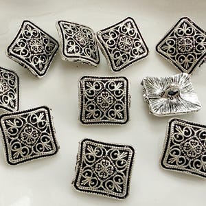 May include: A collection of silver-toned square buttons with an ornate, symmetrical design. The buttons feature a raised, detailed pattern within a square frame, with a dark border. The buttons are arranged on a white surface.