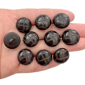 May include: A collection of dark brown, round buttons with a woven texture. One button has a shank, while the others are decorative. The buttons are held in a hand against a white background. These buttons are suitable for sewing and craft projects.