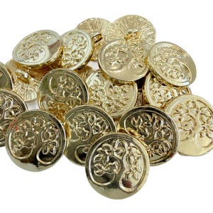 Könnte beinhalten: Eine Ansammlung runder, goldfarbener Metallknöpfe, die jeweils ein erhabenes, geprägtes Motiv eines Baumes mit ausladenden Ästen und Blättern zeigen. Die Knöpfe haben eine polierte, reflektierende Oberfläche und eine kleine Öse auf der Rückseite.