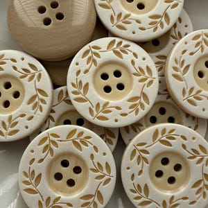 May include: A collection of cream-colored buttons with four holes. Some buttons have a simple design, while others feature a decorative leaf pattern around the center. The buttons are made of a light-colored material.