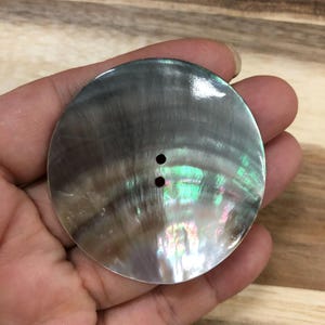 May include: A large, circular, flat button crafted from iridescent mother-of-pearl shell. The surface displays a dark, swirling gray and brown pattern with shimmering highlights of green and pink. It features two small, centered holes for attachment.