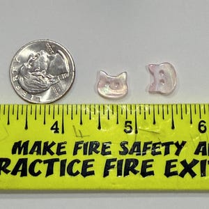 May include: Two iridescent cat-shaped buttons and a US dime are displayed next to a ruler. The buttons are light pink and have a pearlescent sheen. The ruler is yellow with black text that reads "MAKE FIRE SAFETY A PRACTICE FIRE EXIT".