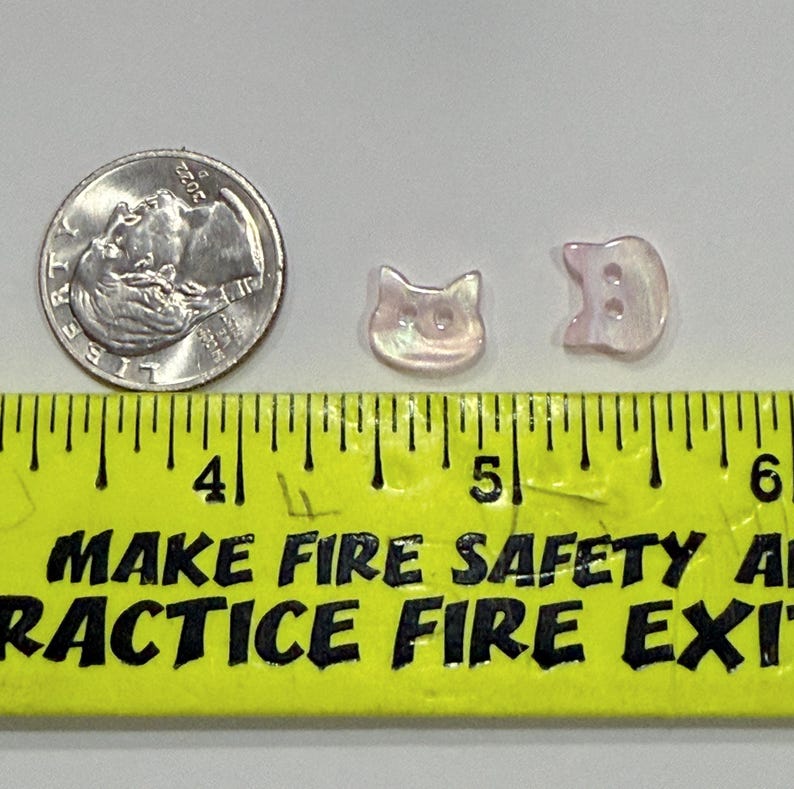 May include: Two iridescent cat-shaped buttons and a US dime are displayed next to a ruler. The buttons are light pink and have a pearlescent sheen. The ruler is yellow with black text that reads "MAKE FIRE SAFETY A PRACTICE FIRE EXIT".