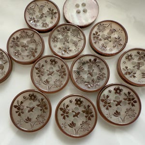 May include: A collection of round buttons with floral designs. The buttons have a light brown border and a clear center with an etched flower pattern. One button has four holes, while the others have two.