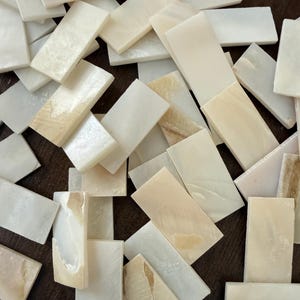 May include: A collection of rectangular mother-of-pearl tiles with a smooth, iridescent finish. Each piece displays natural variations in off-white and cream tones, featuring a flat surface and straight edges, arranged in a scattered, overlapping pile.
