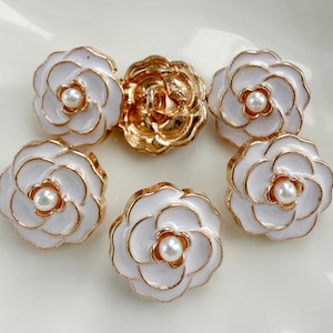 May include: Five round buttons shaped like white camellia flowers with gold-toned metal edges and a small white pearl bead at the center of each. The buttons feature a layered petal design and a metal shank on the back for attachment to fabric.