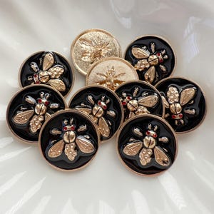 May include: A collection of decorative buttons featuring a bee design. The buttons have a gold-colored bee on a black background. Some bees have red accents. The buttons are round and have a shiny finish.