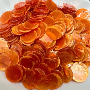 May include: A large collection of circular, translucent orange sequins. Each individual sequin features a smooth, glossy finish and a single small hole positioned near the top edge, designed for sewing or crafting applications.