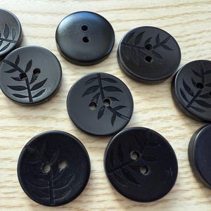 May include: A collection of black buttons with two holes each. Some buttons feature an etched leaf design. The buttons are round and made of a smooth, dark material. They are arranged on a light-colored wooden surface.