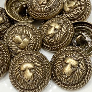 May include: A collection of antique brass-colored buttons. Each button features a detailed lion's head surrounded by a rope-like border. The buttons have a vintage, aged appearance, suggesting they are old or have been designed to look old.