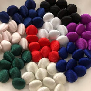 Op de afbeelding: Een verzameling ronde, met stof beklede knopen in diverse kleuren, waaronder rood, wit, zwart, blauw, groen en paars. Elke knoop heeft een gladde, satijnachtige afwerking met een licht bolle vorm, gerangschikt in clusters per kleur.
