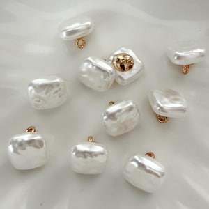 May include: A collection of white, irregular-shaped pearl buttons with gold-colored loops. The buttons have a lustrous, pearlescent finish, and some feature a gold-colored button center. These buttons are ideal for sewing and craft projects.