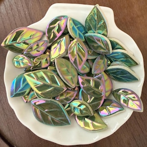 May include: A collection of iridescent ceramic leaf-shaped tiles in shades of green, yellow, and purple, displayed in a white, shell-shaped dish. The leaves have a glossy finish and are arranged in a random pattern.