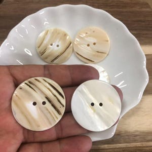 May include: Four round, two-hole buttons with a smooth, polished finish. Two buttons feature a cream and brown marbled pattern, while the other two display a solid, iridescent off-white appearance with subtle natural variations.