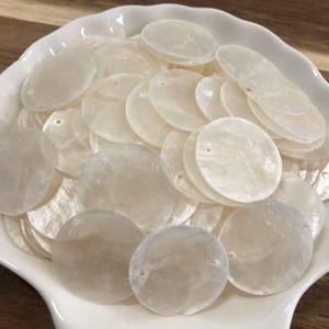 Puede incluir: Una colección de discos circulares de nácar en color blanco roto, cada uno con un pequeño orificio pretaladrado cerca del borde. Estos componentes translúcidos, planos y lisos presentan un brillo iridiscente natural y están apilados.