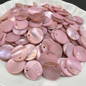 Puede incluir: Una colección de botones redondos de nácar rosa claro, cada uno con un pequeño orificio cerca del borde superior. Estos componentes circulares de concha presentan una superficie lisa e iridiscente con variaciones naturales de tono.