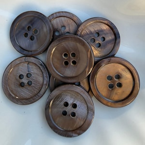 May include: A collection of seven large, round buttons with four holes each. The buttons are a dark brown color with a marbled, iridescent effect. The buttons are arranged on a white surface.