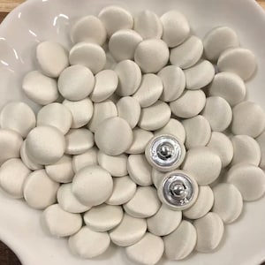 Könnte beinhalten: Eine große Ansammlung runder, mit weißem Stoff bezogener Knöpfe mit einer glatten, satinartigen Oberfläche. Zwei Knöpfe liegen umgedreht, um ihre silberfarbenen Metallösen und die kreisförmigen Rückseiten zu zeigen.