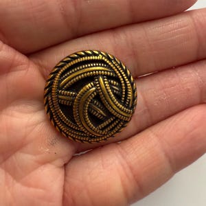 May include: A round, antique brass button with an intricate woven design. The button features a detailed pattern of interwoven lines and a textured edge. The button is approximately 1 inch in diameter and has a vintage aesthetic.