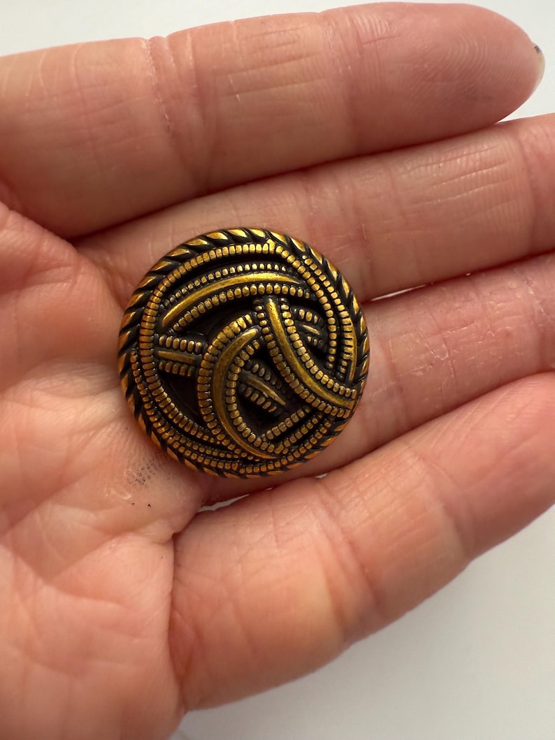 May include: A round, antique brass button with an intricate woven design. The button features a detailed pattern of interwoven lines and a textured edge. The button is approximately 1 inch in diameter and has a vintage aesthetic.