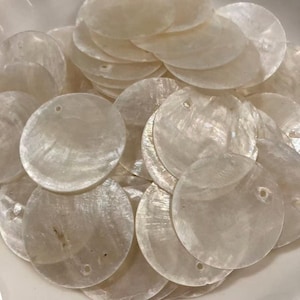 Puede incluir: Una colección de discos circulares de nácar en color blanco roto, cada uno con un pequeño orificio cerca del borde. Los discos tienen una superficie lisa e iridiscente con variaciones naturales en la textura y un aspecto semitranslúcido.