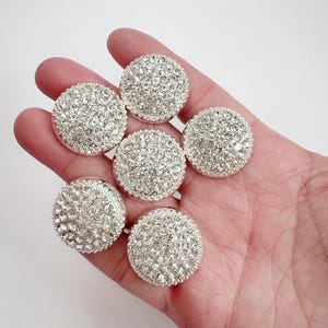 May include: A set of six circular buttons featuring a silver-toned base densely covered in small, clear, faceted crystals. Each button has a rounded, domed profile with a reflective surface that catches light, creating a sparkling appearance.