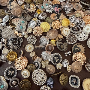 May include: A large collection of assorted decorative buttons in various shapes, sizes, and metallic finishes including gold, silver, bronze, and black. The buttons feature intricate embossed patterns, floral motifs, geometric designs, and embedded faux pearls.