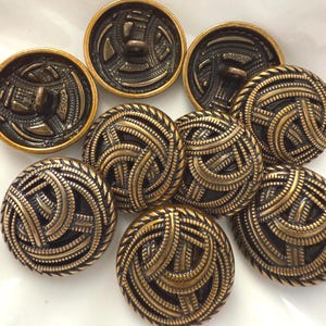 May include: A collection of antique brass buttons with intricate, swirling designs. The buttons have a dark, textured background with raised, golden-colored detailing. The buttons are round and vary in size.